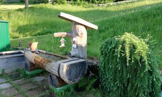 A rustic wooden well stands in a meadow. In the background, green trees and a well-kept lawn can be seen.