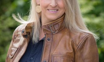 A woman with blonde hair is wearing a brown leather jacket and is smiling. In the background, green plants are visible.