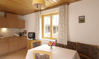 A cozy kitchen with a wooden ceiling, a dining table, and a small living room. Bright curtains and a window create an inviting atmosphere.