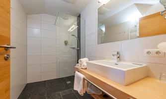 A modern bathroom with a shower and a sink on a wooden shelf. The walls are tiled in light color, and there are towels next to the sink.