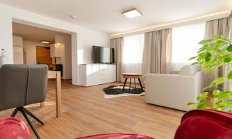 A modern living room with a white sofa, a table, and a television. Large windows let in a lot of light and there is a warm wooden floor.
