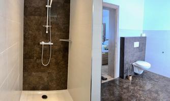 A modern shower with brown tiles and a clear shower wall. In the background, a bathroom with a toilet can be seen.