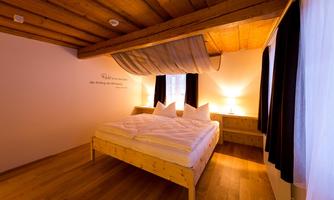 A cozy bedroom with a large bed and wooden ceiling. The room has windows with curtains and a warm atmosphere due to the lighting.