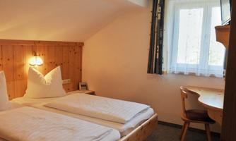 A cozy room with two beds, a small table, and a window with natural light. The walls are in soft colors, and there is wooden paneling.
