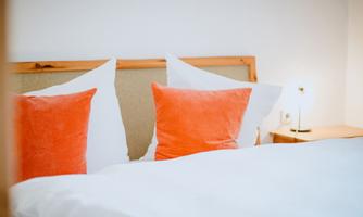 A comfortable bed with white blankets and two orange pillows. Next to the bed is a small lamp on a nightstand.