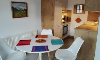 A modern dining room with a round table and white chairs. In the background, a simple kitchenette can be seen.