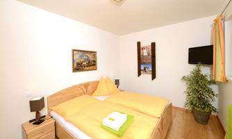 A cozy bedroom with a double bed and yellow bedding. There are pictures hanging on the wall, and there is a TV as well as a houseplant.