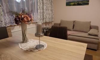 A cozy living area with a dining table, a vase with flowers, and a comfortable sofa. The windows are adorned with curtains and there is a pretty landscape painting on the wall.