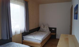 A simple room with two beds and wooden-paneled walls. A window lets in natural light and a desk is positioned against the wall.
