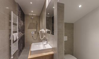 A modern bathroom with a shower, a wooden sink, and a toilet. The walls are in neutral gray tones.