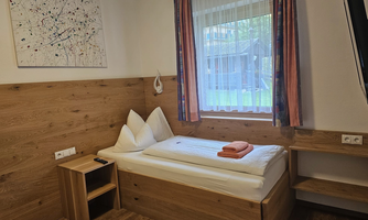 A simple room with a single bed and wood paneling. There is a window with curtains and a small shelf.