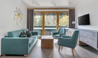 A modern living room with teal sofas and a small wooden table. Large windows offer a view of nature outside.