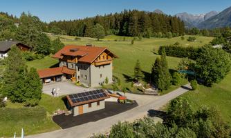 A modern house surrounded by green landscape and trees. In the background, mountains and a clear sky can be seen.