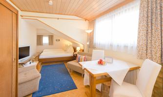 A cozy room with a double bed, a television, and a small dining area. The room has large windows and bright wood decor.