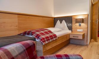 A cozy bedroom with a single bed and wooden paneling on the walls. The bed linen features modern red-and-white check patterns.