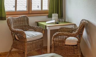 A cozy room with two rattan chairs and a small table. The windows are framed by green curtains.