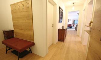 A bright hallway with wooden floors and white walls. Coat hooks hang on the wall and a bench stands on one side.