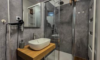 A modern bathroom with a shower cubicle and an elegant wash basin on a wooden shelf. The walls are in a plain gray tone and the room appears well organized.