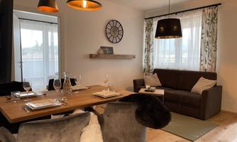 A modern dining room with a wood-stained table and elegant chairs. In the background, there is a cozy sofa and large windows that provide plenty of light.