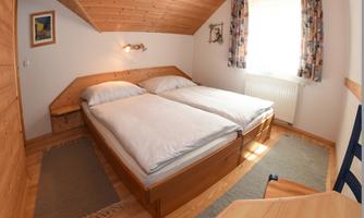 A cozy bedroom with two single beds and wooden paneling. The room has a window with curtains and a small table.
