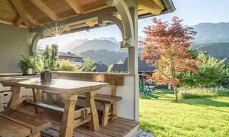 A cozy terrace with a wooden table and benches, surrounded by a green meadow. In the background, mountains and a beautiful tree can be seen.