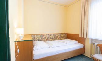 A simple room with a single bed and a small bedside table. The walls are painted light yellow and the window lets in plenty of daylight.