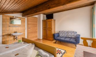 A cozy bedroom with a double bed, a blue sofa, and wooden flooring. The walls are bright, and there is a wardrobe as well as a small washbasin.