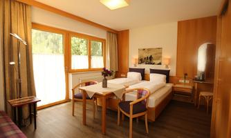 A bright bedroom with two single beds, wooden floor, and large windows. There is a table with chairs and decorative elements.