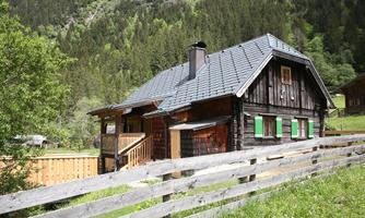 A cozy wooden house with a black roof and green shutters.  
Surrounded by a picturesque landscape with mountains and trees.
