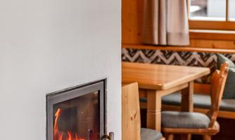 A cozy living room area with a wood stove and a dining table. The room has wooden paneling and windows with curtains.