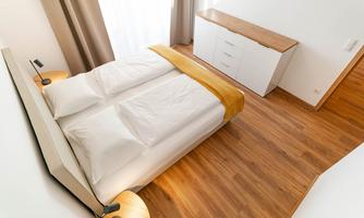 A modern bedroom with a large bed and a wooden sideboard. The room has bright walls and a warm wooden floor.