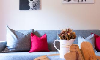 A cozy living room with a gray couch and red pillows. On a table are wooden hearts and a vase with dried flowers.