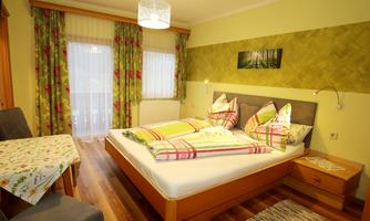 A cozy bedroom with a large bed and colorful pillows. The room has a bright atmosphere and a view of the outdoors through the windows.