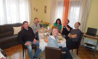 A group of six people sits at a table in a living room. There is food and drinks on the table, and the atmosphere feels relaxed and friendly.