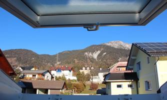 View through an open window of a picturesque village with houses and green meadows. In the background, majestic mountains rise under a clear blue sky.