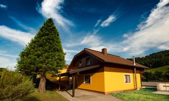 A yellow house with a beautiful roof and large windows stands against a clear blue sky. A large tree and green grass surround the building.