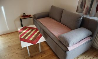 A cozy living room with a gray sofa and a small table. On the table lies a decorative placemat in red and gold.