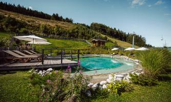 A beautiful natural pool surrounded by green grass and colorful flowers. In the background, trees and a gentle range of hills can be seen.