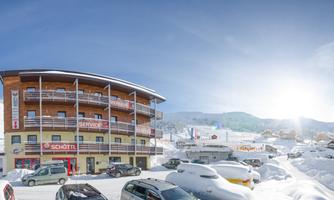 A cozy building in a snowy winter landscape with many cars in the parking lot. The sun is shining brightly in the blue sky.