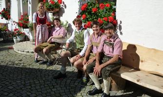 A group of five people in traditional Bavarian costumes sits comfortably on a bench. In the background, red geraniums bloom and the weather is sunny.