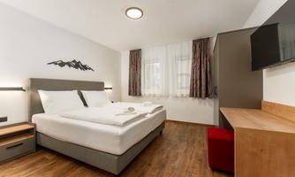 A modern hotel room with a large double bed and wooden flooring. There is a desk, a television, and large windows with curtains.