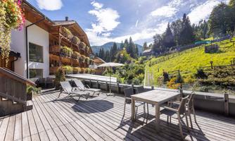 A bright terrace with wooden flooring and modern loungers. In the background, green meadows and mountain landscapes can be seen.