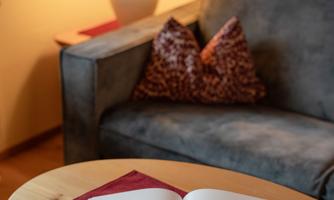A cozy living space with an open book on a table. In the background, there is a lamp and a sofa with cushions.