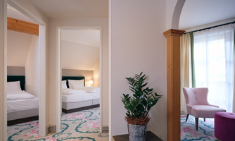 A stylish room with a view of another sleeping area. The rooms are bright and modernly furnished, with a beautiful carpet and a plant in the foreground.