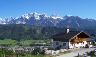 A picturesque mountain landscape with snow-covered peaks and a green valley. In the foreground stands a traditional wooden house.