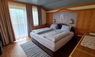 A cozy bedroom with a large bed and light wood interior. Gentle rays of light fall onto the carpet in front of the windows.