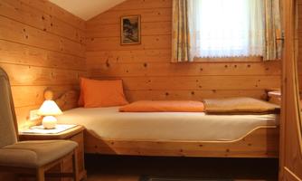 A cozy bedroom with wooden walls and natural light. There is a bed, a chair, and a table lamp.