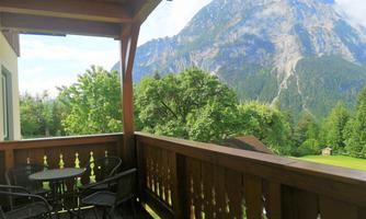 A balcony with two chairs and a table offers a view of majestic mountains and lush trees. The clear air and peaceful surroundings create a relaxing atmosphere.
