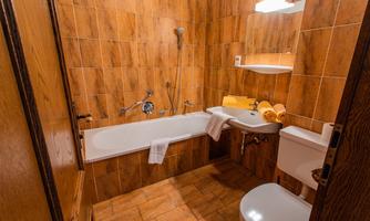 A small bathroom with brown tiles and white bathroom furniture. There is a bathtub, a sink, and towels on a shelf.