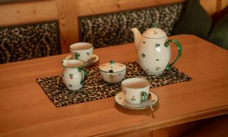 An elegant teapot with matching cups sits on a wooden table. The dinnerware is adorned with delicate green patterns.